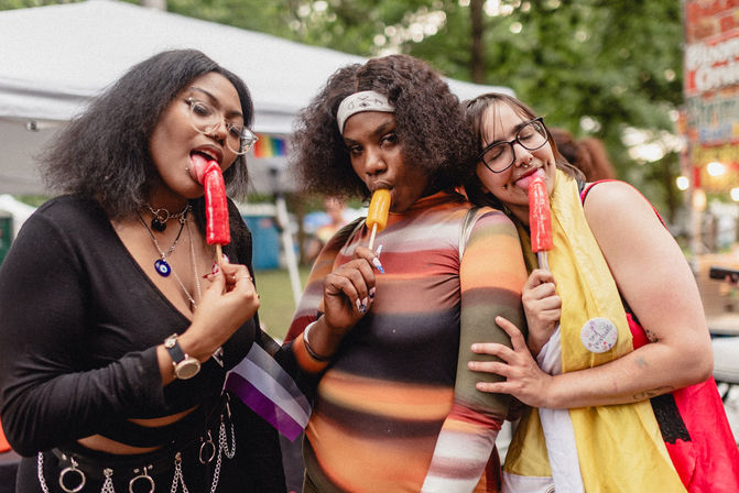 Three people at an outdoor Pride festival enjoying colorful popsicles, posing under park tents in playful outfits with pins and small flags.