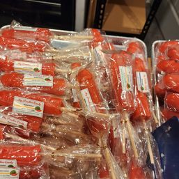 Tray of bright red watermelon-flavored candy pops on wooden sticks, individually wrapped in clear plastic and stacked in a market or candy shop display.