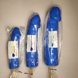 Three blue novelty candy lollipops shaped like penises, small to large sizes, individually wrapped in clear plastic with labels (blue raspberry), laid on a light background