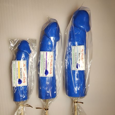 Three blue novelty candy lollipops shaped like penises, small to large sizes, individually wrapped in clear plastic with labels (blue raspberry), laid on a light background