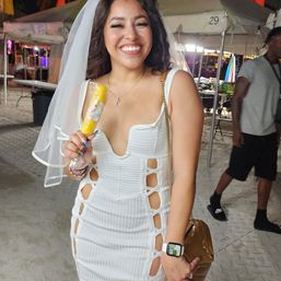 Smiling woman in a white cut-out dress and veil holding a yellow popsicle at a nighttime outdoor tented beach festival