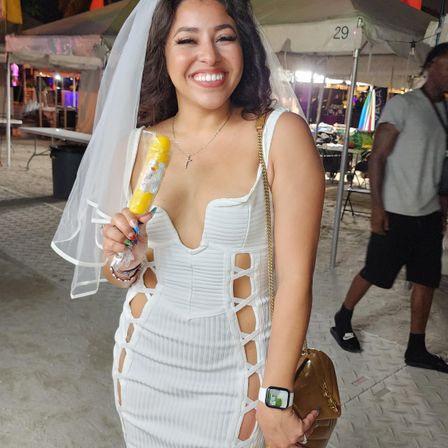 Smiling woman in a white cut-out dress and veil holding a yellow popsicle at a nighttime outdoor tented beach festival