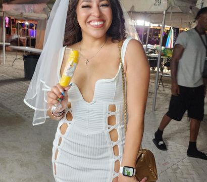 Smiling woman in a white cut-out dress and veil holding a yellow popsicle at a nighttime outdoor tented beach festival