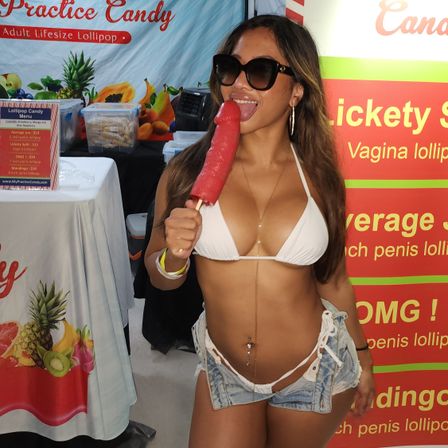 Woman in white bikini and sunglasses at a festival vendor booth playfully licking a large red novelty adult lollipop, wearing denim shorts and a belly chain.