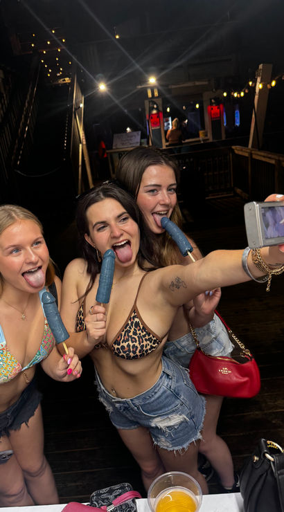 Three friends in bikinis take a selfie on a wooden boardwalk at night, licking blue popsicles under string lights — playful summer beach nightlife vibe with drinks and party energy.