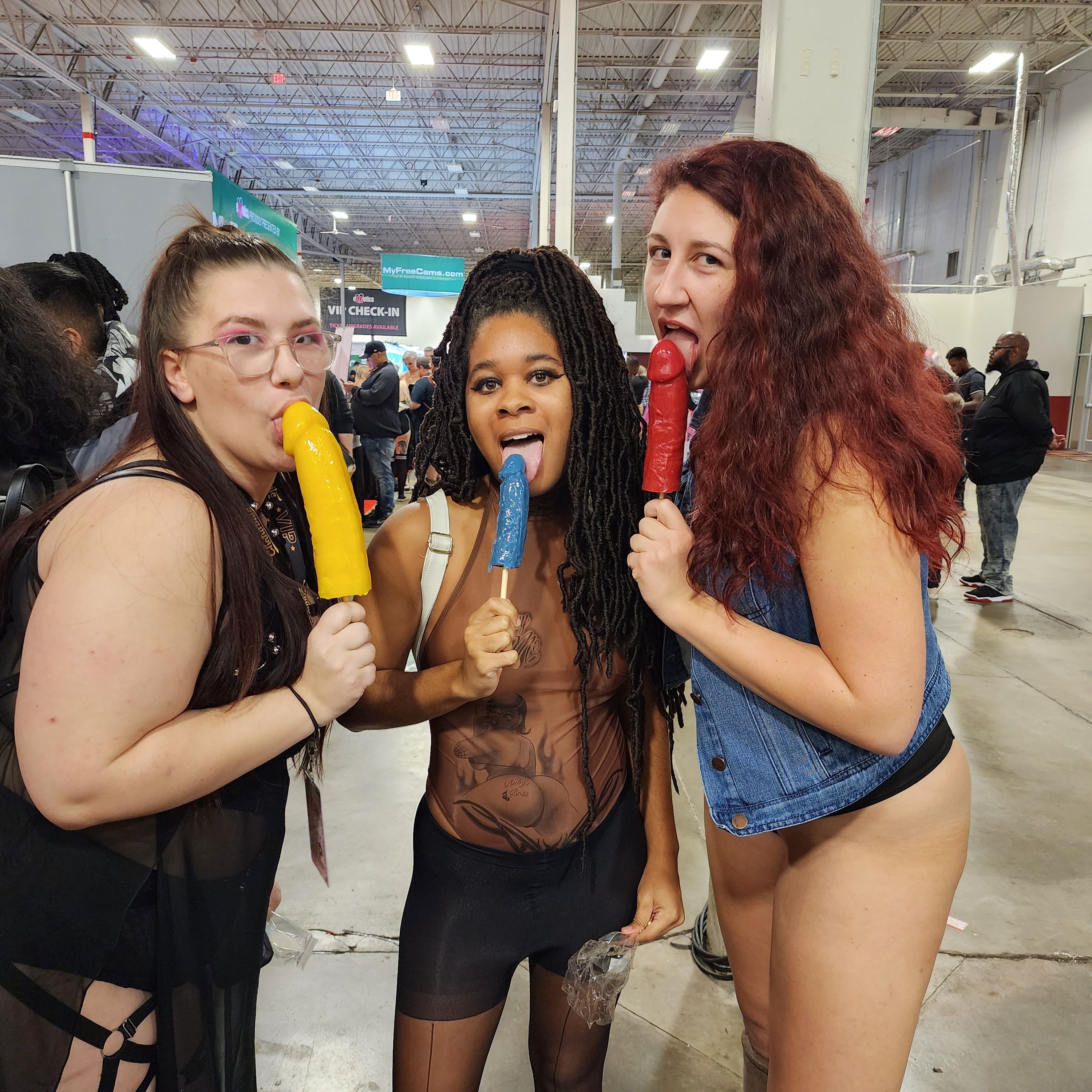 Three adults in playful outfits at an indoor expo hall playfully licking colorful phallic novelty popsicles (yellow, blue, red) amid a busy convention crowd.