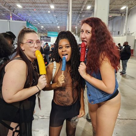 Three adults in playful outfits at an indoor expo hall playfully licking colorful phallic novelty popsicles (yellow, blue, red) amid a busy convention crowd.
