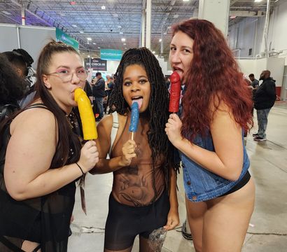 Three adults in playful outfits at an indoor expo hall playfully licking colorful phallic novelty popsicles (yellow, blue, red) amid a busy convention crowd.