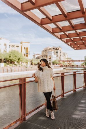 Woman in a white cable-knit turtleneck sweater, black leggings and snakeskin ankle boots leaning on a rust-colored railing along an urban canal promenade with mixed-use buildings under a sunny sky