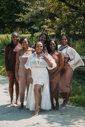 Joyful group of six women posing barefoot on a sunlit park path, wearing coordinated neutral-toned outfits; the center woman in white shows a ring as friends smile against a leafy green backdrop.