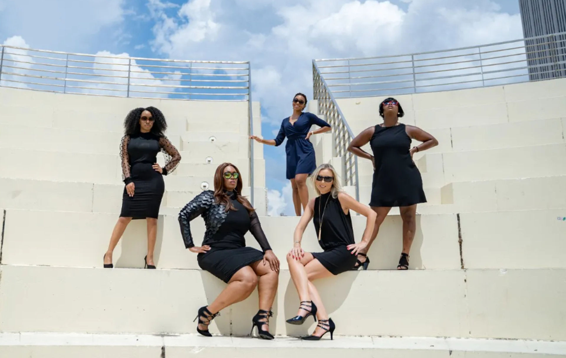 Five women in stylish black dresses and heels pose confidently on white concrete rooftop steps with metal railings, city skyscrapers and a bright blue sky with fluffy clouds in the background.