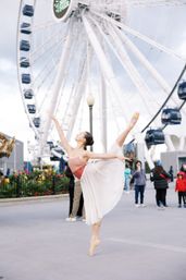 Ballet dancer on pointe in a flowing skirt and pink leotard, striking an arabesque in front of a large waterfront Ferris wheel at an urban pier, with onlookers and flowers nearby.
