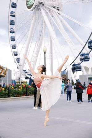 Ballet dancer on pointe in a flowing skirt and pink leotard, striking an arabesque in front of a large waterfront Ferris wheel at an urban pier, with onlookers and flowers nearby.