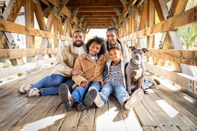 Smiling family portrait of parents and two children with their dog seated on a wooden covered bridge in a sunlit autumn park