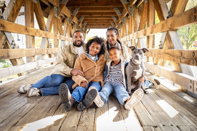 Smiling family portrait of parents and two children with their dog seated on a wooden covered bridge in a sunlit autumn park
