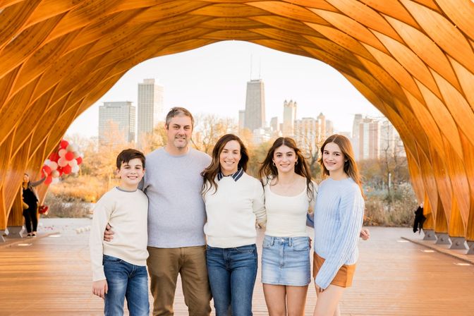 Smiling family of five posing under a curved wooden pavilion with the Chicago skyline in the background and red balloons to the side