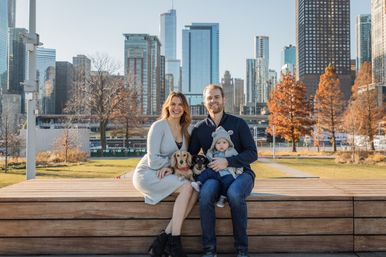 Smiling family enjoying autumn in a downtown waterfront park—parents on a wooden bench with a baby in a hooded jacket and two small dogs, with orange trees and glass skyscrapers of the city skyline behind them.