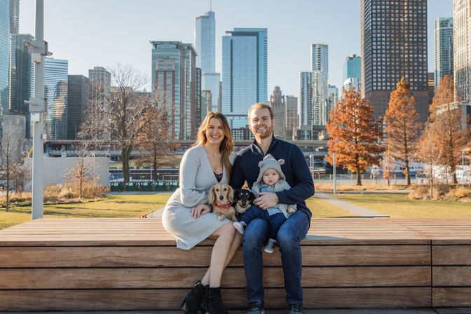 Smiling family enjoying autumn in a downtown waterfront park—parents on a wooden bench with a baby in a hooded jacket and two small dogs, with orange trees and glass skyscrapers of the city skyline behind them.