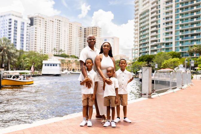 Family of five in coordinated white and beige outfits posing on a sunny waterfront boardwalk by a marina with boats and high‑rise condo skyline
