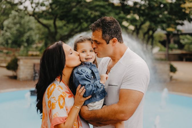 Outdoor family portrait: parents kiss their smiling young daughter in a denim jacket while dad holds her in front of a park fountain and leafy trees.
