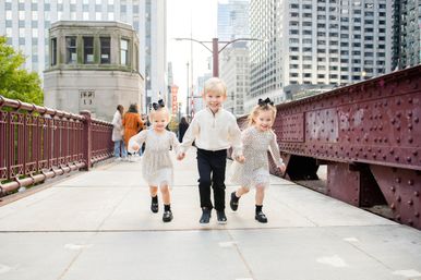 Three happy children — a boy in a white sweater and two girls in polka-dot dresses with black bows — holding hands and running across a red iron bridge in downtown Chicago with skyscrapers in the background.