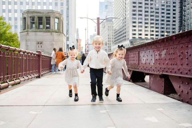 Three happy children — a boy in a white sweater and two girls in polka-dot dresses with black bows — holding hands and running across a red iron bridge in downtown Chicago with skyscrapers in the background.