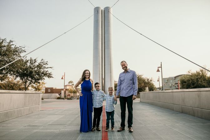 Smiling family of four (parents and two young boys) standing on a modern cable-stayed pedestrian bridge in a downtown urban plaza at dusk