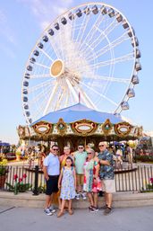Smiling multigenerational family posing in front of a giant Ferris wheel and colorful carousel at a sunny waterfront amusement park.