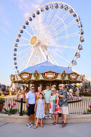 Smiling multigenerational family posing in front of a giant Ferris wheel and colorful carousel at a sunny waterfront amusement park.