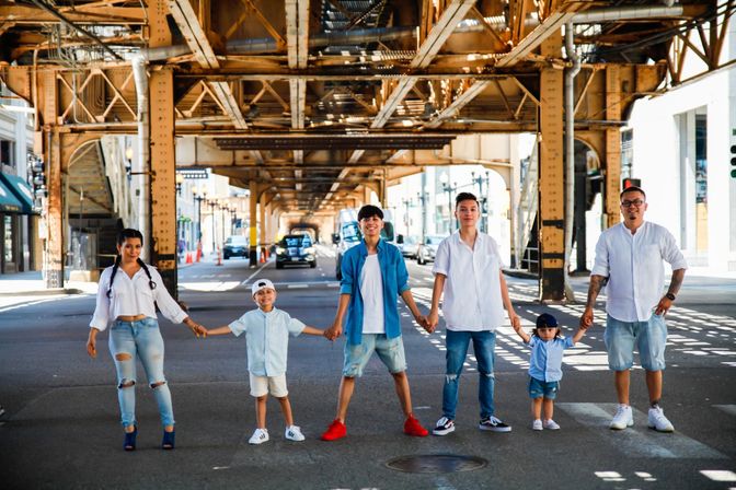Family of six holding hands across a sunlit city street under rusted elevated train tracks, casual summer outfits in a playful urban photoshoot
