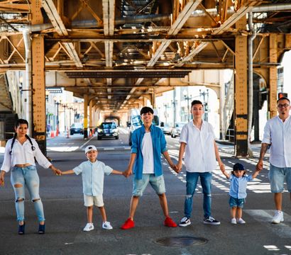 Family of six holding hands across a sunlit city street under rusted elevated train tracks, casual summer outfits in a playful urban photoshoot