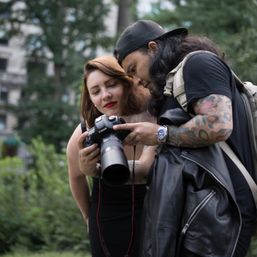 Two photographers checking images on a Canon DSLR in a leafy city park — woman with red lipstick and a man with a tattooed arm and backpack reviewing the camera screen.