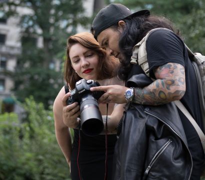Two photographers checking images on a Canon DSLR in a leafy city park — woman with red lipstick and a man with a tattooed arm and backpack reviewing the camera screen.
