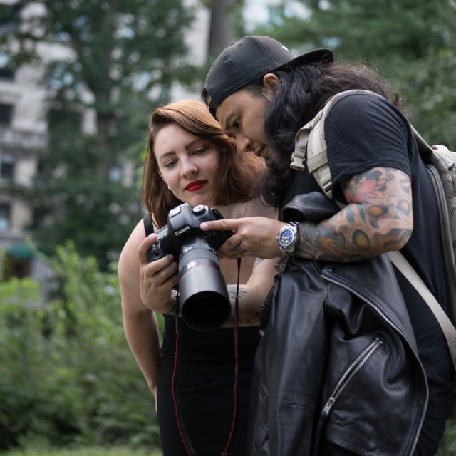 Two photographers checking images on a Canon DSLR in a leafy city park — woman with red lipstick and a man with a tattooed arm and backpack reviewing the camera screen.