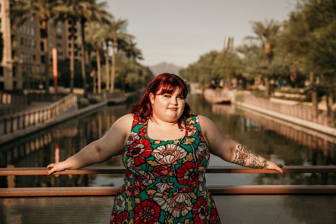 Person in a vibrant floral dress leaning on a railing by an urban canal lined with palm trees and buildings at golden hour, tattooed arm visible and distant mountains reflected in the water.