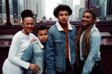 Four smiling people posing on a steel city bridge with a blurred downtown skyline; cozy sherpa denim jackets, a white sweater, and long braids for a casual urban family portrait.