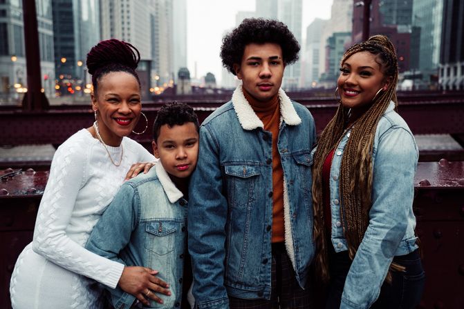 Four smiling people posing on a steel city bridge with a blurred downtown skyline; cozy sherpa denim jackets, a white sweater, and long braids for a casual urban family portrait.