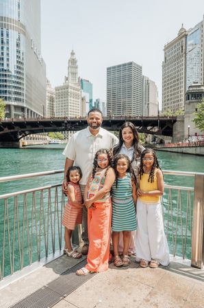 Sunny family of six posing at the Chicago Riverwalk railing, children in colorful summer outfits with downtown skyscrapers and a bridge in the background.