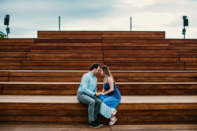 Romantic couple sitting on wide wooden amphitheater steps outdoors, holding hands and touching foreheads; woman in a blue sundress and man in a light blue shirt.