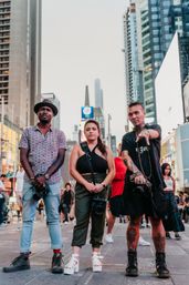 Three street photographers posing in Times Square, New York City — a diverse trio with cameras against a backdrop of skyscrapers and bright billboards.