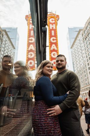 Couple embracing on a downtown sidewalk beside a reflective storefront, framed by a tall red 'Chicago' theater marquee and surrounding skyscrapers.
