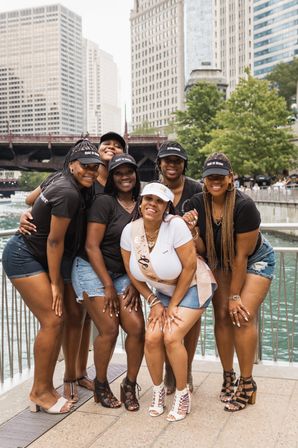 Bachelorette group of six women on the Chicago Riverwalk — bride-to-be in a white sash and visor with friends in matching black shirts and caps, city skyline and bridge in the background.