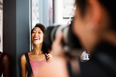 Laughing young woman posing for a photographer on a city sidewalk, camera lens blurred in the close foreground