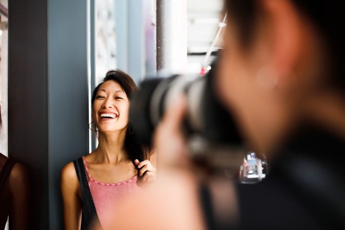 Laughing young woman posing for a photographer on a city sidewalk, camera lens blurred in the close foreground