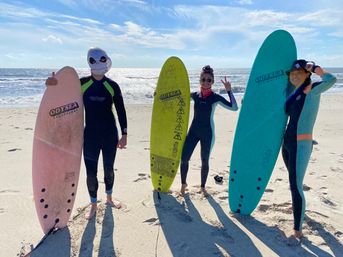 Three surfers in wetsuits holding pink, yellow and teal surfboards on a sunny sandy beach with sparkling ocean and blue sky behind, one wearing a skull mask and another flashing a peace sign.