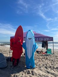 Sunny beach scene with two people in inflatable costumes — a blue shark and red lobster holding a red surfboard — in front of a purple canopy tent and ocean waves.