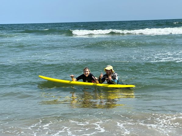 Two smiling surfers on a bright yellow surfboard in shallow ocean waves near the beach, flashing shaka signs under a clear blue sky.
