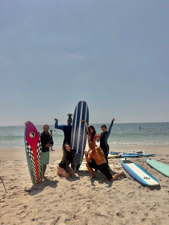 Group of six surfers posing with colorful surfboards on a sunny sandy ocean beach, smiling and celebrating after a session.