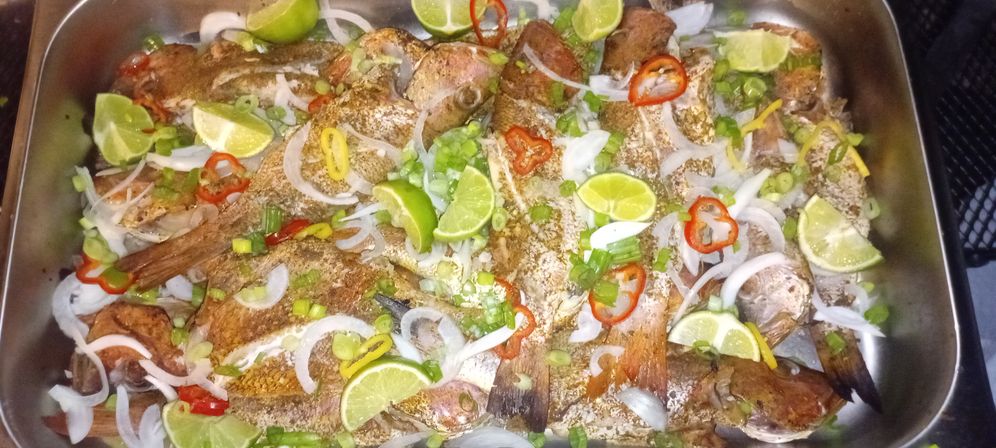 Tray of whole spiced fried fish garnished with lime wedges, sliced onions, scallions and red and yellow chili rings — zesty seafood platter.