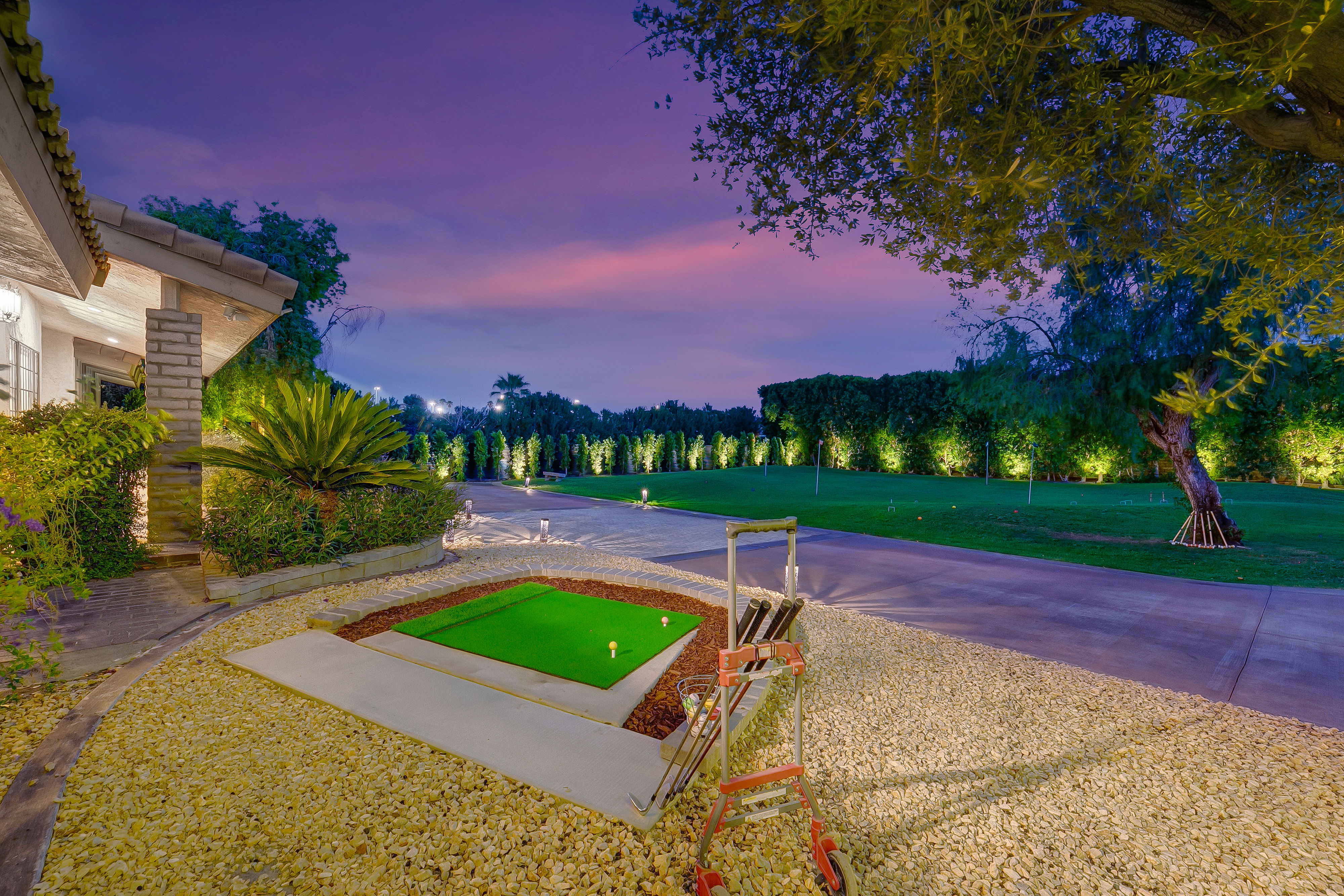 Well-lit residential backyard putting green and practice tee with golf clubs on a push cart, decorative stone landscaping and illuminated trees under a purple twilight sky.